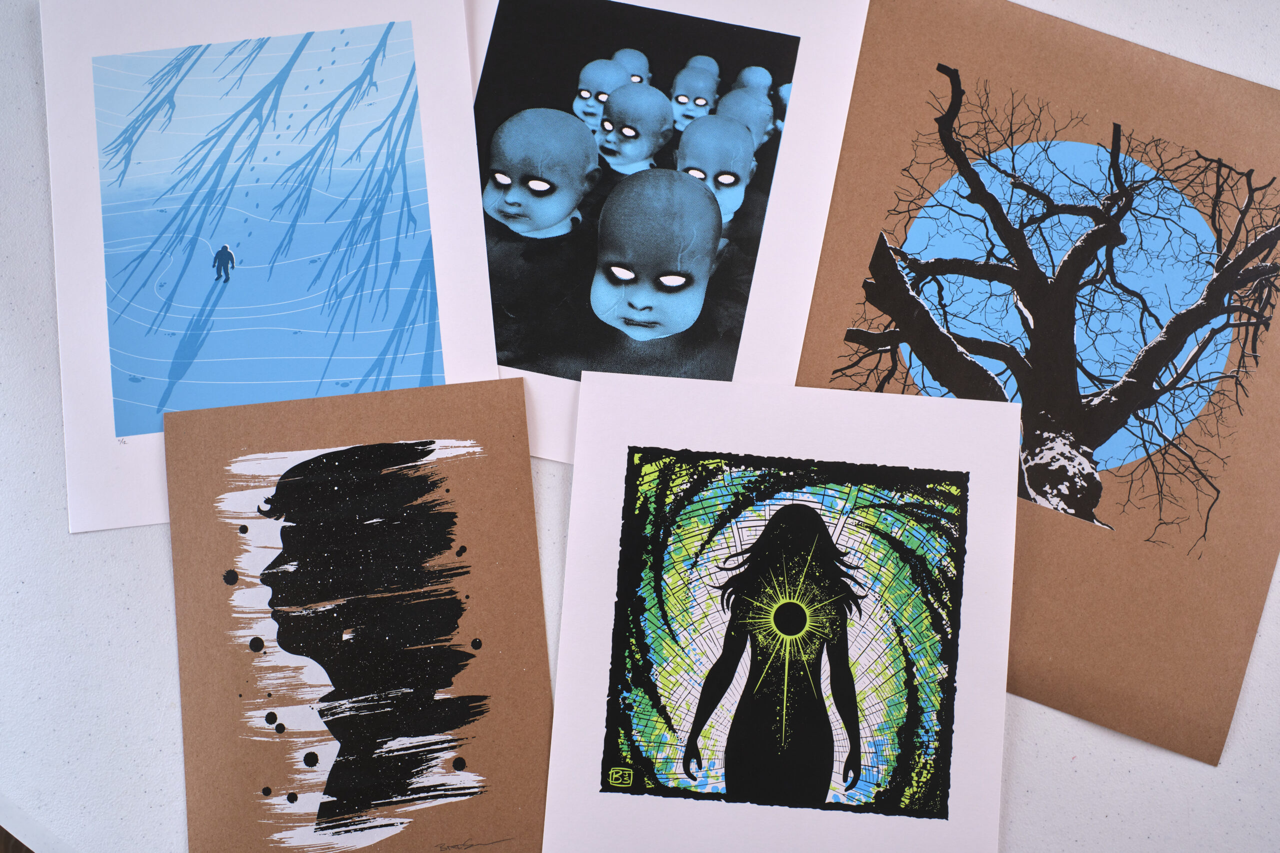 A collection of screen printed posters and artwork, including a man on a frozen lake, spooky halloween decorations, a tree silhouetted by a blue moon, a brush stroke portait profile, and a comic art superheroine